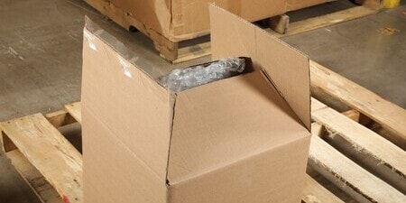 Close-up of an open cardboard box on a wooden pallet, showing tape peeling off the flaps.
