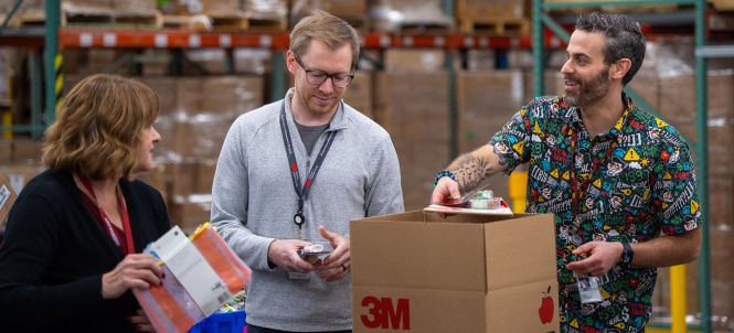 Three employees with blurred faces stand around a 3M-branded box in a warehouse, one sealing the box while another holds colorful folders.
