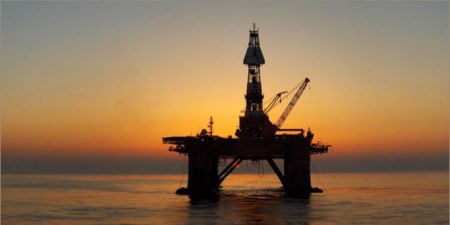 Offshore oil platform silhouetted against a sunset over calm ocean waters
