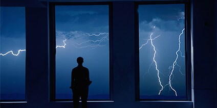 Photo of man standing in front of windows looking at lighting storm at night
