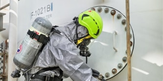 Worker in full hazmat suit and breathing apparatus inspecting an industrial tank for safety compliance.
