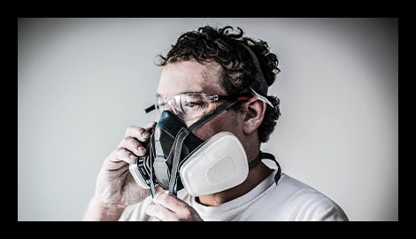 Worker fitting a 3M reusable half-facepiece respirator with safety glasses, shown in close-up profile.