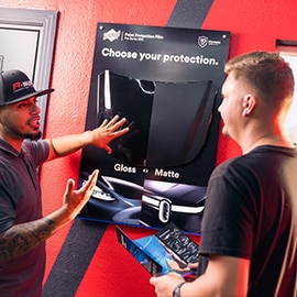 Two technicians review a large 3M marketing display in a shop, illustrating dealer support for consumer leads.
