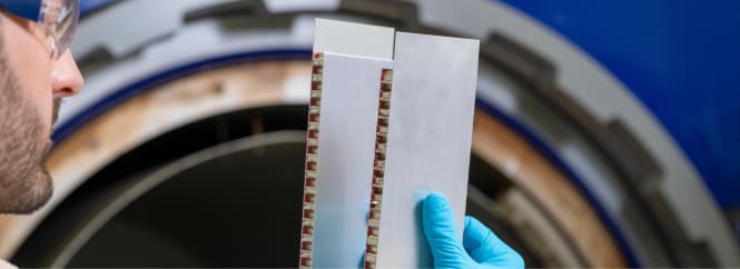 Researcher in safety glasses and blue glove holds a layered test sample in front of lab equipment, inspecting material structure and alignment.
