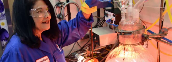 A scientist wearing a blue lab coat and yellow gloves is working with a large glass apparatus in a laboratory.
