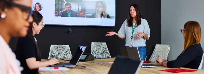 Woman presents in a conference room beside a screen showing remote participants, while colleagues seated at a table work on laptops and take notes.
