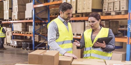 Two warehouse workers in high-visibility vests discuss logistics while holding a tablet in a large distribution center.
