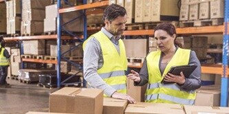 Two warehouse workers in high-visibility vests discuss logistics while holding a tablet in a large distribution center.
