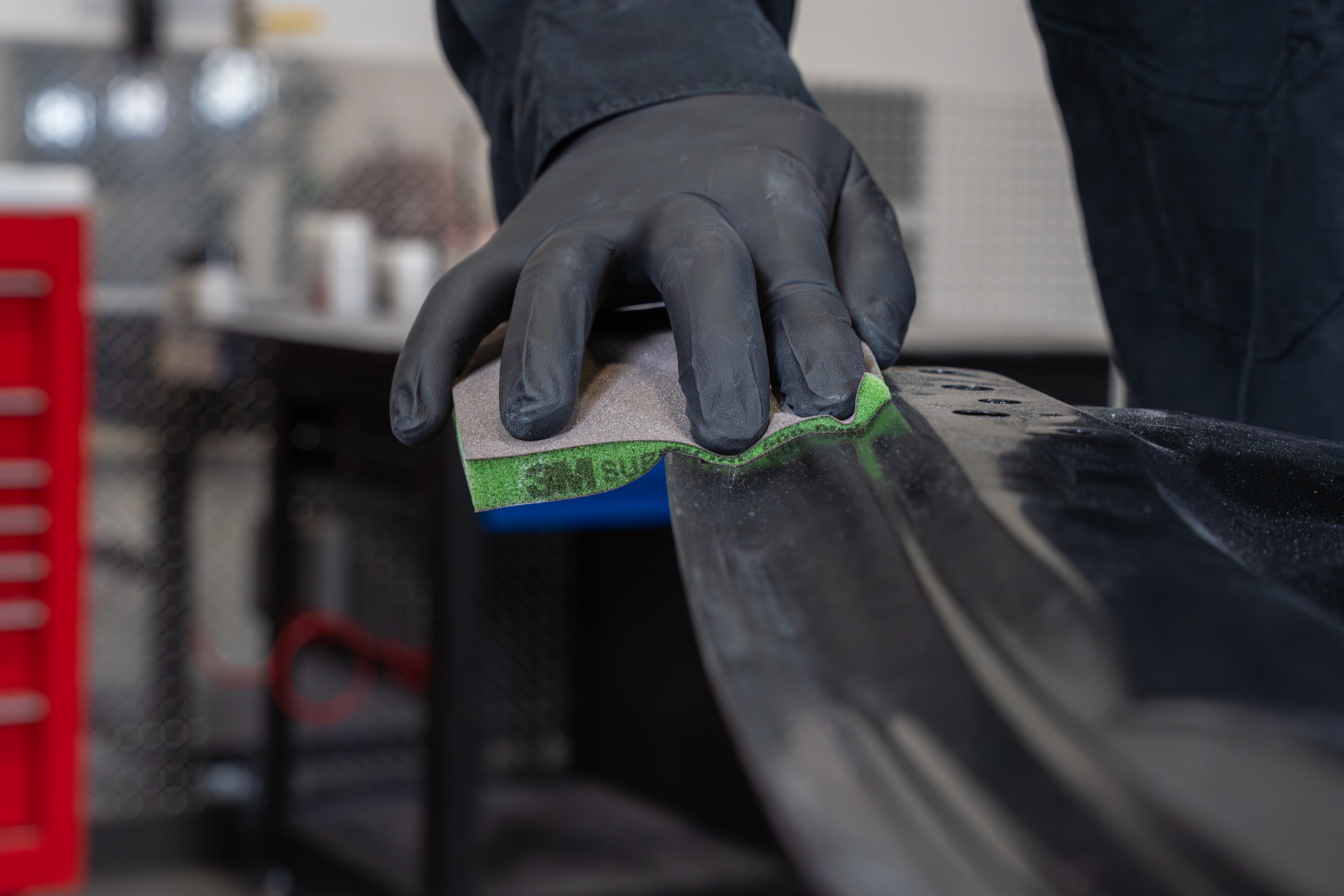 Close-up of a gloved hand using an abrasive sanding pad on an automotive surface in a workshop.
