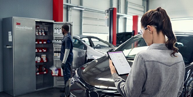 A person standing next to a vehicle in an auto body shop looks at the screen of a tablet device. A shop employee on the other side of the vehicle looks inside of a : 3M™ RepairStack™ cabinet.
