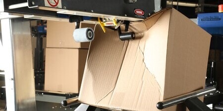 A cardboard box being crushed by an industrial packaging machine, illustrating structural stress.
