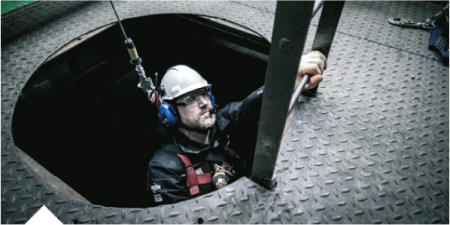 Worker wearing safety gear climbs a ladder out of a confined space while attached to a fall-protection system.
