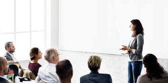 A woman leads a business presentation in front of a large white screen for an audience
