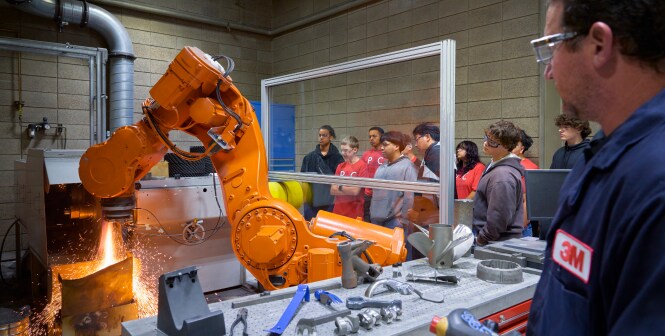 Orange industrial robot arm welding metal behind a safety screen as students in red shirts and staff watch in a factory training space with tools in foreground.
