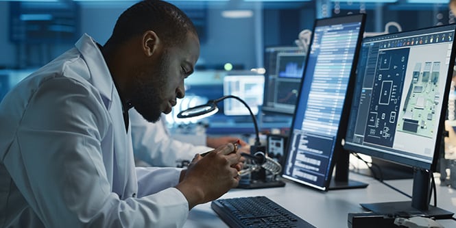 A person in a lab coat works at a desk with multiple monitors displaying technical interfaces in an industrial workspace.
