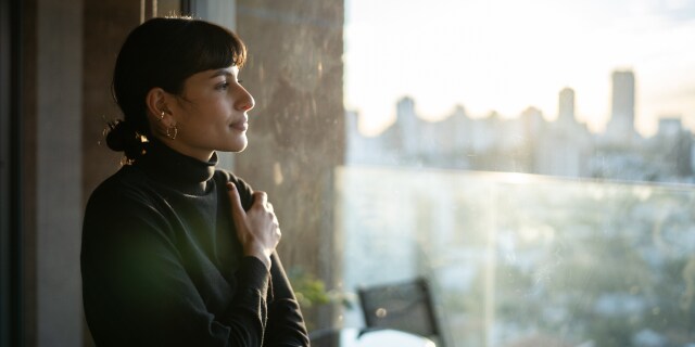 Person standing beside a window overlooking a city skyline.
