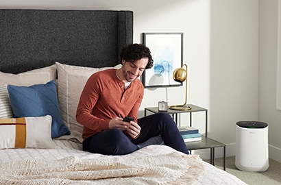 A man sitting on bed next to a Filtrete&trade; Smart Air Purifier.