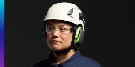 Person wearing a white safety helmet with attached 3M Peltor earmuffs against a dark background.
