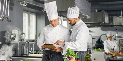 Two chefs confer over a tablet in a commercial kitchen.
