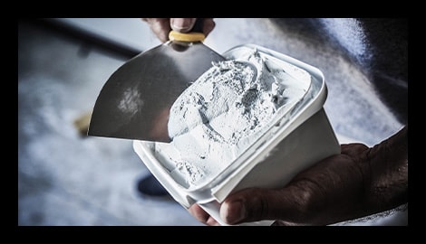 Hands loading a wide putty knife with white wall spackle from a plastic container.