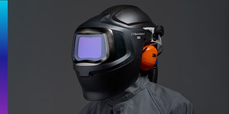 Person wearing a black 3M Speedglas welding helmet with attached orange earmuffs and a powered air system hose.
