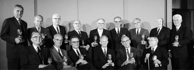 Black-and-white group portrait of 14 suited men at an awards event, arranged in rows, many holding statuettes, against a plain indoor backdrop.
