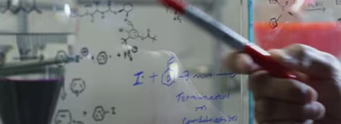 A person is writing chemical equations and molecular structures on a transparent board using a red marker.
