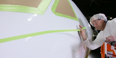 Image of a worker applying masking tape to the nose of an aircraft whose cockpit windows are already masked
