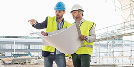 Two construction managers in hard hats, with one pointing and one holding construction blueprints.