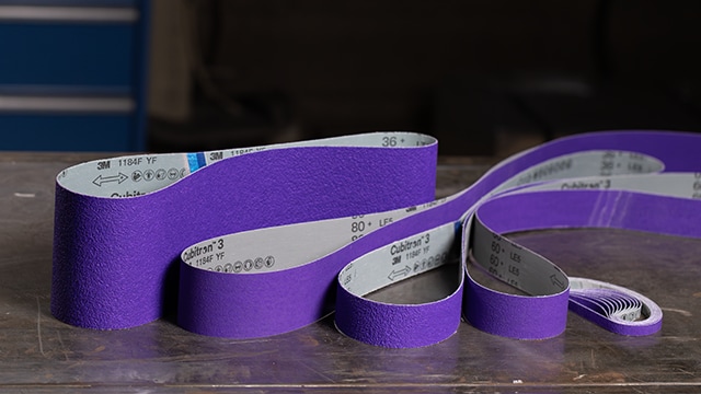 Purple 3M abrasive belts arranged on a workbench in a workshop setting.

