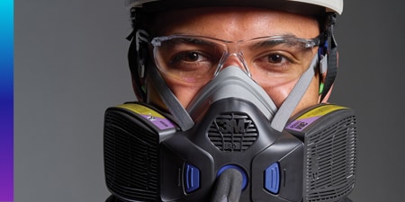Close‑up of a person wearing clear safety glasses and a 3M half‑face respirator with dual filters.
