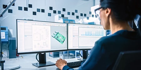 Engineer reviewing 3D PCB designs on dual monitors in a design workspace.
