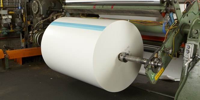 A large roll of white paper with a blue splicing tape stripe on a heavy-duty industrial manufacturing press.
