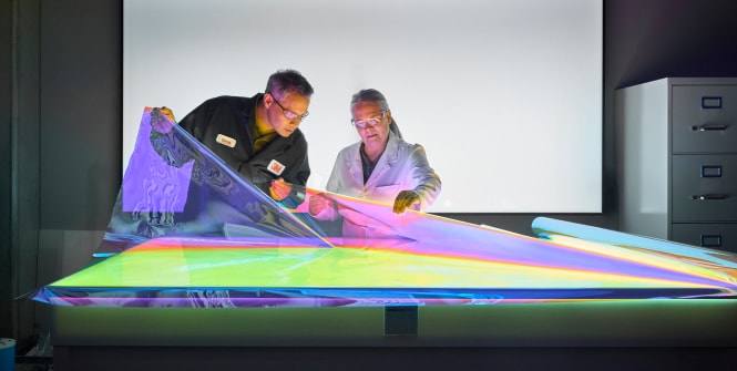 Two 3M employees examine a large iridescent film sheet under colorful lighting in a lab or demonstration setting.
