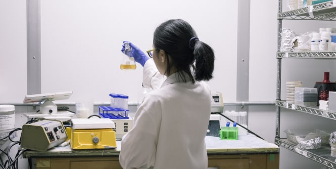 A scientist wearing a lab coat and gloves is working in a laboratory, holding a container with a yellow liquid.
