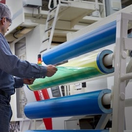 A technician in a hairnet and safety glasses handles a roll of green mounting tape on a storage rack.