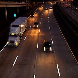 Cars and a semi-truck illuminate a four-lane highway and pavement markings with their headlights