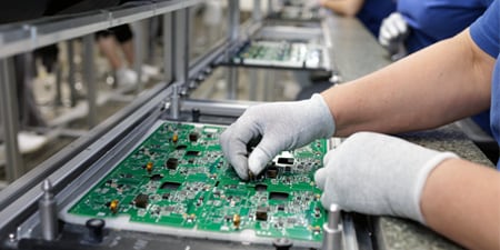 Gloved workers assembling and inspecting green PCB boards on a production line.

