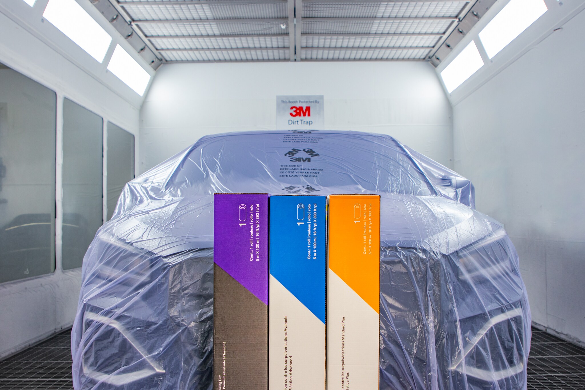 Three boxes of 3M™ Plastic Sheeting stand in front of a vehicle covered with clear protective plastic inside an automotive paint booth.
