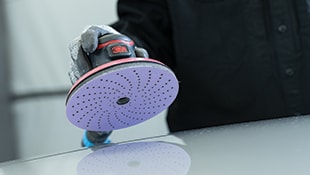 A gloved hand holding a 3M orbital sander with a perforated sanding disc against a smooth surface.
