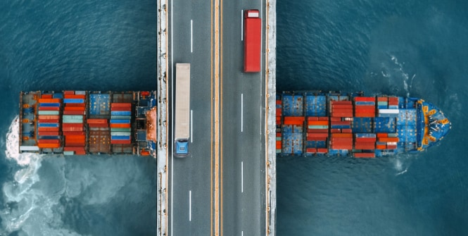 Overhead view of a blue truck crossing a bridge above a container ship loaded with red and blue cargo containers on the water.
