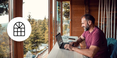 Person working on a laptop beside large windows, with window icon overlay. 
