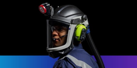 Worker wearing a protective helmet with a face shield, mounted headlamp, earmuffs, and a powered air‑purifying respirator hose, shown in profile against a dark background.
