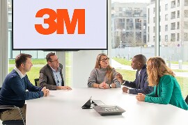 A group of five people sitting around a conference table in a modern office setting, and a large screen displaying the 3M logo in the background.