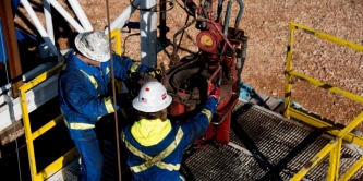 Two industrial workers in protective clothing operating heavy equipment on an elevated work platform at a job site.
