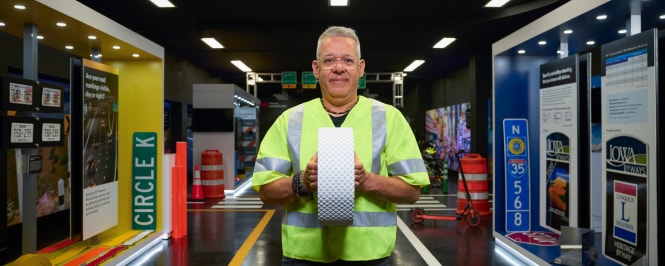 Worker in a high-visibility safety shirt holds a white textured sample block in an indoor exhibit or training space with signage and displays in the background.
