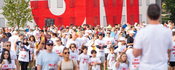 Crowd of people in white 3M shirts gathered outdoors at a community event, with a speaker in the foreground and a large red 3M backdrop behind them.
