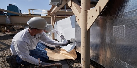 Two people wearing hard hats and gloves apply a roll of insulating material to siding.
