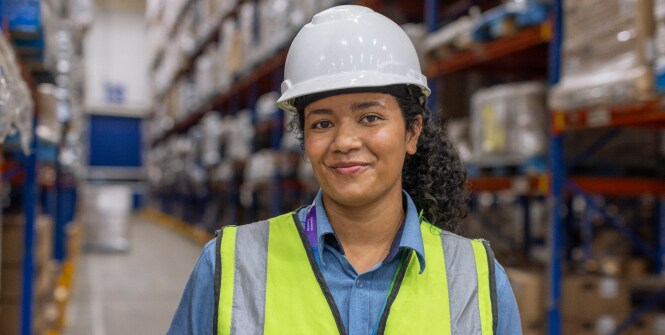 Warehouse worker wearing a hard hat and safety vest, with face blurred for privacy, standing in front of shelves stocked with materials.
