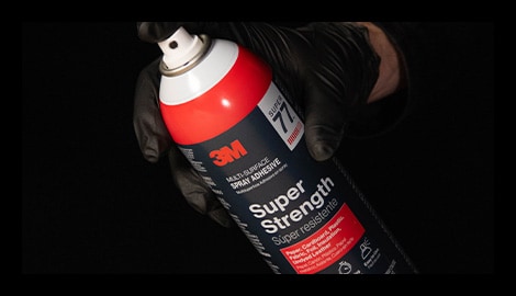 Gloved hand holding a can of 3M Super Strength Spray Adhesive against a black background.